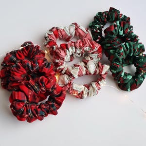 May include: A collection of fabric scrunchies in various patterns. The scrunchies feature red and green plaid, red and white striped, and green and red patterned fabrics. The scrunchies are arranged on a white surface with a string of fairy lights.