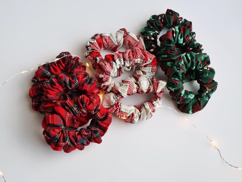 May include: A collection of fabric scrunchies in various patterns. The scrunchies feature red and green plaid, red and white striped, and green and red patterned fabrics. The scrunchies are arranged on a white surface with a string of fairy lights.