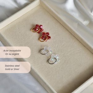 K&ouml;nnte beinhalten: Zwei Paar Creolen mit blumenf&ouml;rmigen Anh&auml;ngern. Ein Paar hat rote Glitzerblumen an goldfarbenen Ringen, das andere Paar hat irisierende wei&szlig;e Blumen an silberfarbenen Ringen. Der Text auf dem Bild lautet Acier Inoxydable Or ou argent und Stainless steel Gold or Silver.