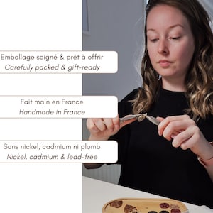 K&ouml;nnte beinhalten: Eine Metallzange wird verwendet, um einen kleinen Metallanh&auml;nger zusammenzusetzen. Drei Textfelder sind sichtbar: Emballage soign&eacute; & pr&ecirc;t &agrave; offrir, Fait main en France und Sans nickel, cadmium ni plomb, jeweils mit englischer &Uuml;bersetzung.