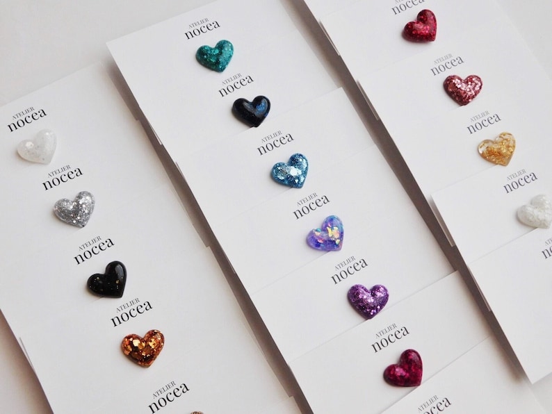 May include: A collection of heart-shaped embellishments in various colors, including teal, black, silver, gold, and purple, are displayed on white cards. Each card is labeled with the text "ATELIER nocea". The hearts have a glittery or textured appearance.