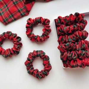 May include: A collection of red, green, and yellow plaid scrunchies. Some scrunchies are scattered on a white surface, while others are neatly arranged in a white box. A matching plaid fabric is also visible.
