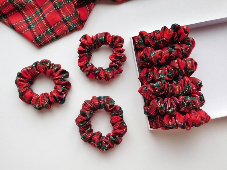 May include: A collection of red, green, and yellow plaid scrunchies. Some scrunchies are scattered on a white surface, while others are neatly arranged in a white box. A matching plaid fabric is also visible.