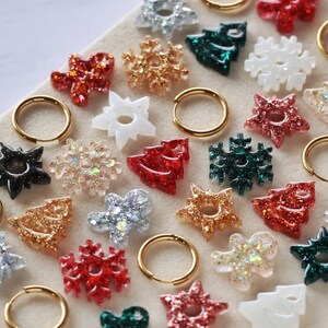Peut inclure: Un assortiment de boucles d'oreilles et de breloques festives en forme de flocons de neige, d'étoiles et de sapins de Noël. Les boucles d'oreilles sont des créoles dorées. Les breloques sont pailletées et de couleurs rouge, vert, or et argent.