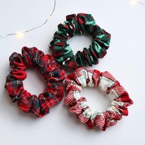 May include: Three fabric scrunchies in festive holiday patterns. One is red and green plaid, another is red and white with green accents, and the third is red and green with a holly design. The scrunchies are on a white surface with string lights.