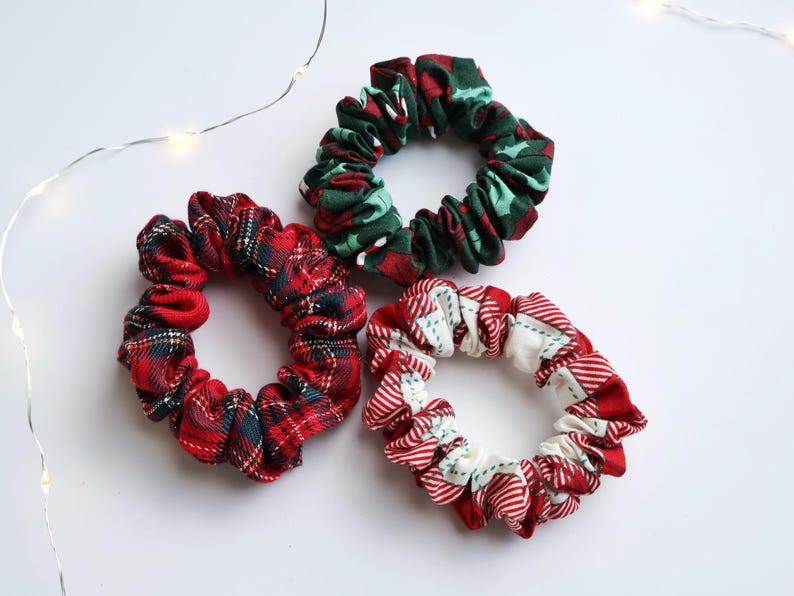 May include: Three fabric scrunchies in festive holiday patterns. One is red and green plaid, another is red and white with green accents, and the third is red and green with a holly design. The scrunchies are on a white surface with string lights.