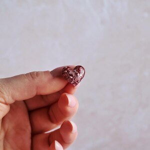 May include: A small, heart-shaped ornament with a glittery, reddish-brown surface. The heart is held between two fingers against a soft, neutral background. The glitter catches the light, creating a sparkling effect. The ornament is a symbol of love and affection.