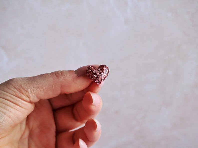 May include: A small, heart-shaped ornament with a glittery, reddish-brown surface. The heart is held between two fingers against a soft, neutral background. The glitter catches the light, creating a sparkling effect. The ornament is a symbol of love and affection.
