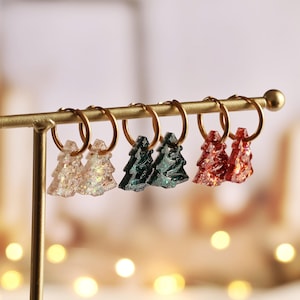 Puede incluir: Tres pares de pendientes de aro con pequeños dijes brillantes en forma de árbol. Los dijes son de color blanco translúcido, verde oscuro y rojo, suspendidos de finos aros de metal dorado en un soporte de exhibición de metal dorado.
