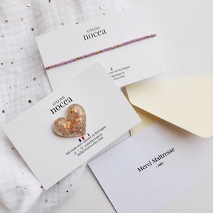 May include: A delicate lavender beaded bracelet with gold accents rests on a white card. A heart-shaped, glitter-filled charm is displayed on another card. The cards are arranged with cream-colored envelopes on a white background with gold specks.
