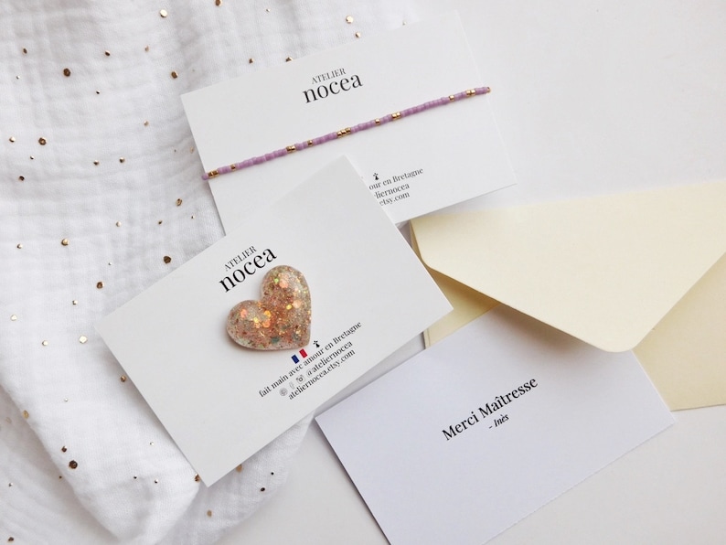 May include: A delicate lavender beaded bracelet with gold accents rests on a white card. A heart-shaped, glitter-filled charm is displayed on another card. The cards are arranged with cream-colored envelopes on a white background with gold specks.