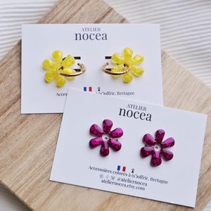 Könnte beinhalten: Zwei Paar blumenförmige Ohrringe auf weißen Präsentationskarten mit der Aufschrift Atelier nocea. Ein Paar hat leuchtend gelbe Blütenblätter an goldenen Creolen, das andere Paar besteht aus dunkelmagentafarbenen Blüten mit Glitzereffekt.