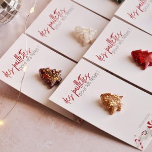 Könnte beinhalten: Nahaufnahme mehrerer weihnachtsbaumförmiger Ornamente mit Glitzer. Die Ornamente sind in verschiedenen Farben, darunter Gold, Rot und Silber, und werden auf weißen Karten mit rotem Text mit der Aufschrift "des paillettes pour les fêtes!" präsentiert.