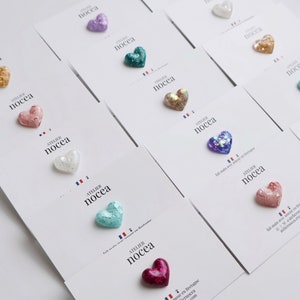 May include: A collection of heart-shaped embellishments in various colors, including gold, pink, purple, and teal, are displayed on white cards. Each heart has a glittery texture. The cards feature the text "ATELIER NOCECA" and a French flag.
