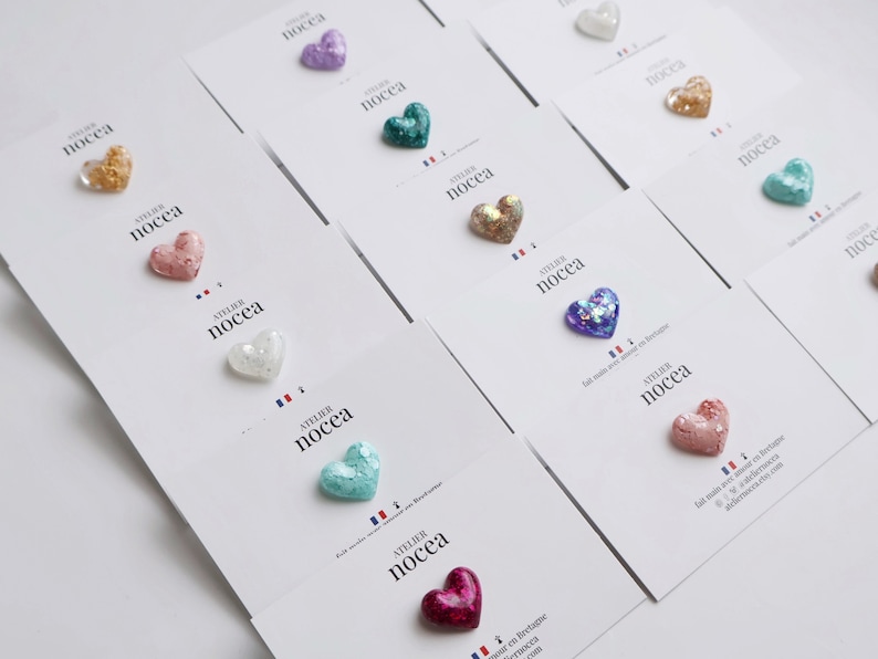 May include: A collection of heart-shaped embellishments in various colors, including gold, pink, purple, and teal, are displayed on white cards. Each heart has a glittery texture. The cards feature the text "ATELIER NOCECA" and a French flag.
