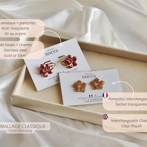 K&ouml;nnte beinhalten: Zwei Paar Creolen aus Edelstahl mit austauschbaren blumenf&ouml;rmigen Anh&auml;ngern auf wei&szlig;en Pr&auml;sentationskarten mit der Aufschrift Atelier Nocea. Ein Paar hat rote glitzernde Blumenanh&auml;nger, das andere Paar hat goldene glitzernde Blumenanh&auml;nger.