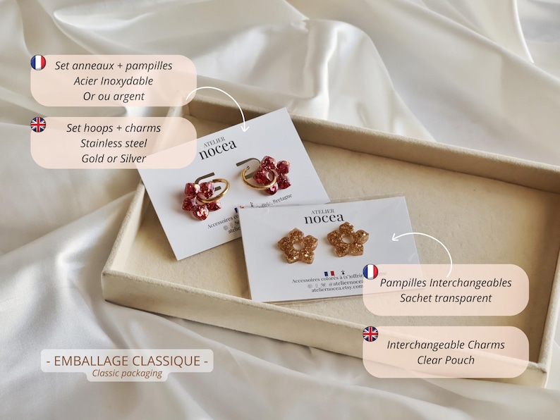 K&ouml;nnte beinhalten: Zwei Paar Creolen aus Edelstahl mit austauschbaren blumenf&ouml;rmigen Anh&auml;ngern auf wei&szlig;en Pr&auml;sentationskarten mit der Aufschrift Atelier Nocea. Ein Paar hat rote glitzernde Blumenanh&auml;nger, das andere Paar hat goldene glitzernde Blumenanh&auml;nger.