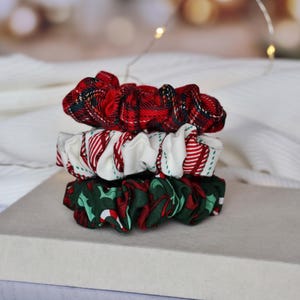May include: Three festive scrunchies stacked on a white surface. The top scrunchie is red plaid, the middle one is white with red stripes, and the bottom one is green with red holly leaves. The background is blurred with soft lighting.