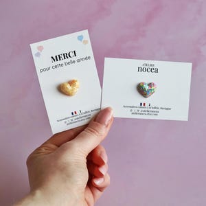 May include: Two heart-shaped pins on white cards are held against a pink background. One pin is gold-colored, and the other is multicolored. The cards have text in French and the brand name "Atelier Nocea".