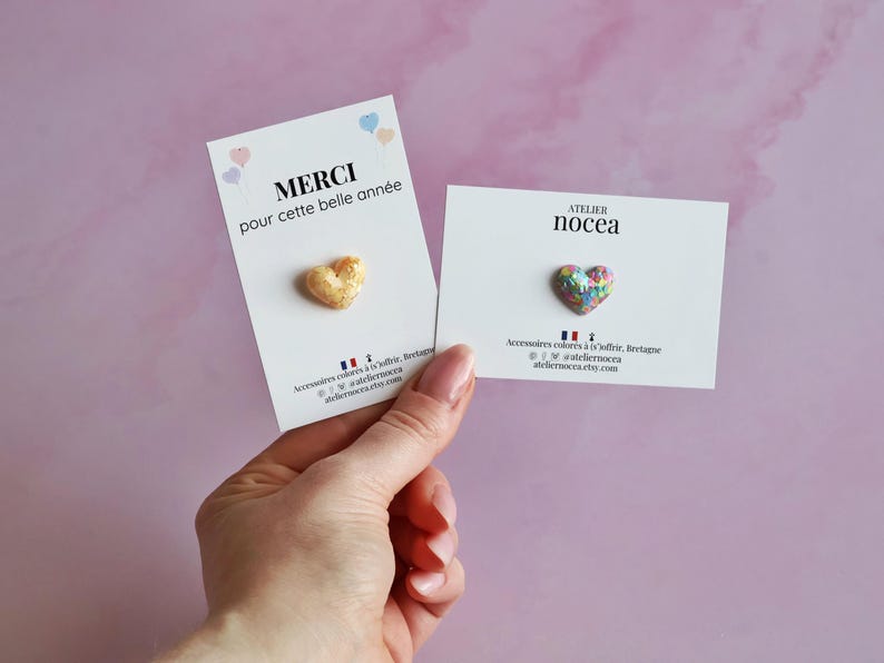 May include: Two heart-shaped pins on white cards are held against a pink background. One pin is gold-colored, and the other is multicolored. The cards have text in French and the brand name "Atelier Nocea".