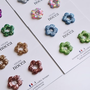 Può includere: Una collezione di fermagli per capelli a forma di fiore con finiture glitterate in vari colori tra cui viola, verde, oro, blu e rosa. I fermagli sono fissati su cartoncini bianchi con il testo Atelier nocea e le informazioni di contatto.