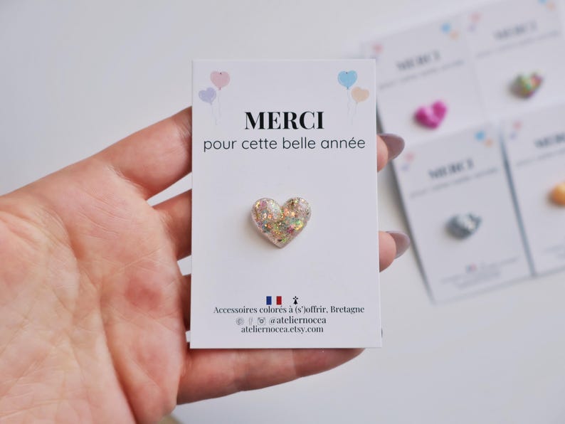 May include: A heart-shaped pin with glitter and iridescent accents is displayed on a white card. The card has the French word "MERCI" and the phrase "pour cette belle ann&eacute;e" printed on it. Other cards with pins are visible in the background.