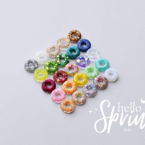 May include: A collection of colorful, glittery donut-shaped beads in various colors, including silver, gold, blue, green, and pink. The words "hello Spring 2026" are written in white script on the bottom right corner. The beads are arranged on a white surface.