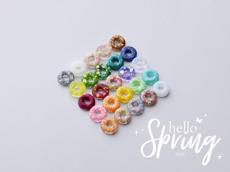 May include: A collection of colorful, glittery donut-shaped beads in various colors, including silver, gold, blue, green, and pink. The words "hello Spring 2026" are written in white script on the bottom right corner. The beads are arranged on a white surface.