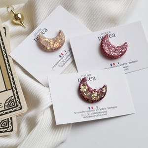 Könnte beinhalten: Drei halbmondförmige Broschen mit Glitzer in Rosa-, Gold- und Brauntönen, präsentiert auf weißen Karten. Die Karten tragen Text und eine französische Flagge. Ein goldfarbenes Pendel und Tarotkarten sind ebenfalls zu sehen.