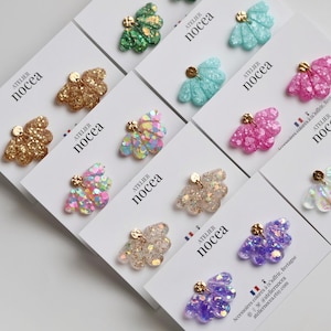 Può includere: Una collezione di orecchini a bottone glitterati e colorati in vari colori, tra cui oro, verde, rosa e viola, esposti su cartoncini bianchi. Ogni orecchino ha un perno color oro e il marchio "ATELIER Nocéa" è stampato sui cartoncini.