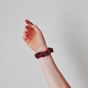 May include: A red and black plaid scrunchie is worn on a wrist. The scrunchie is made of a soft fabric and has a gathered design. The background is a plain, off-white color.
