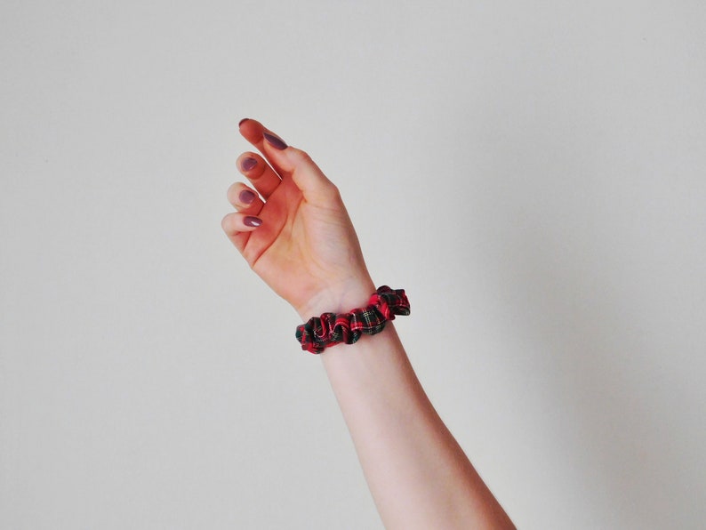May include: A red and black plaid scrunchie is worn on a wrist. The scrunchie is made of a soft fabric and has a gathered design. The background is a plain, off-white color.