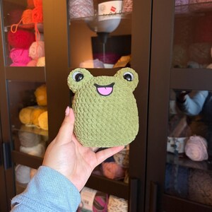 May include: A green crocheted frog plushie with black and white eyes and a pink mouth. The frog is held in front of a dark brown cabinet with shelves filled with yarn in various colors.
