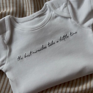 Puede incluir: Body de bebé blanco con mangas cortas y cuello redondo. El texto "the best miracles take a little time" está impreso en escritura negra en la parte delantera. El body está sobre una tela a rayas beige y blanca.