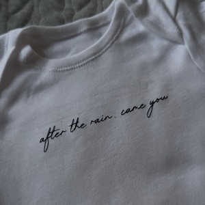 May include: White baby onesie with the phrase "after the rain, came you" in black script. The onesie has a round neckline and short sleeves. The fabric appears to be soft and comfortable.