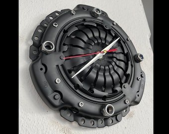 May include: A black metal clock with a unique design featuring a circular clutch plate with exposed bolts. The clock hands are silver and red.