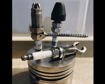 May include: A silver metal container with three spark plugs attached to it. The spark plugs are connected by a metal screw and a wrench. The container is sitting on a wooden surface.