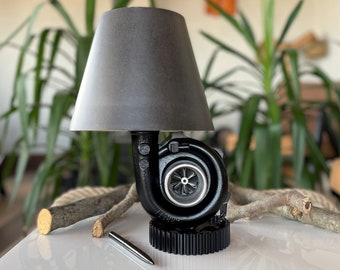 May include: A black lamp with a gray lampshade, made from a repurposed car turbocharger. The lamp is sitting on a white surface with a pen and two pieces of driftwood.