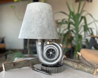 May include: A gray lampshade with a silver turbocharger base. The turbocharger is mounted on a gear base. The lamp is on a white table with a pen and a branch.