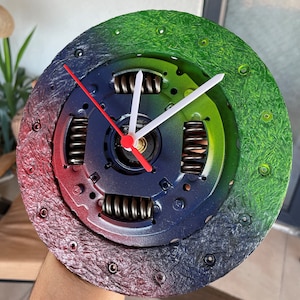 May include: A colorful clock with a green, blue, and red design. The clock face is a car part, possibly a clutch plate, with a black background and silver metal parts. The clock hands are white and red.