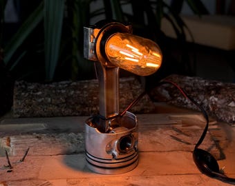 Puede incluir: Una lámpara de escritorio única hecha de piezas de motor recicladas. La lámpara presenta una base de pistón plateada, una biela y una cálida bombilla de estilo Edison. La lámpara está cableada con un cable y un enchufe negros y se coloca sobre una superficie de madera.