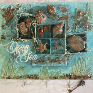 May include: A handcrafted wall art piece with a coastal theme. It features a light blue background with embedded seashells, starfish, and other marine elements. The phrase "Down by the Sea" is written in white script. Includes hooks for keys.