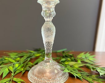 May include: A clear glass candle holder with a decorative etched design. The holder has a wide base and a tall, slender stem.