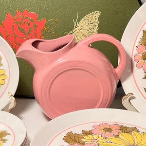 May include: A pink ceramic pitcher with a curved handle and spout, set against a backdrop of floral-decorated plates. The plates feature a white base with a pink rim and a floral pattern in yellow, pink, and brown. A decorative butterfly is visible.