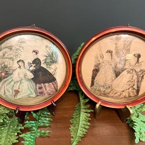 May include: Two circular wall frames with reddish-brown wooden borders, each containing a vintage-style illustration. The left frame shows two figures in period clothing outdoors, while the right frame depicts two figures in gowns indoors.