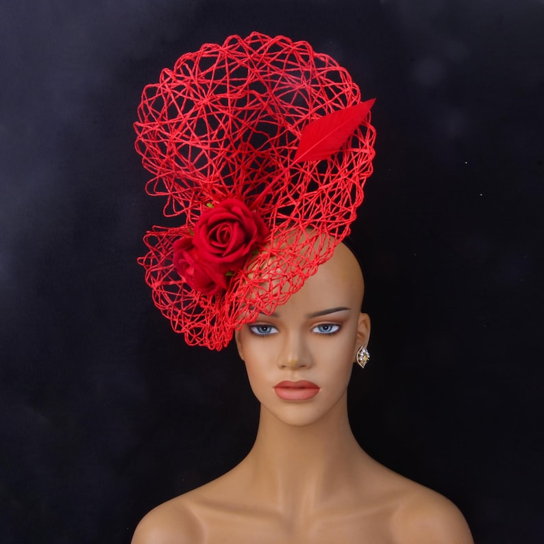 May include: A bright red fascinator hat featuring an intricate, open-weave wire structure. The headpiece is adorned with two prominent red fabric roses and a single matching red feather, creating a bold, sculptural design that sits to one side.