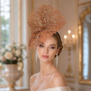 Puede incluir: Un tocado fascinator de color cobre con una intrincada estructura de alambre en forma de red, sujeta a una diadema oscura. El tocado presenta un diseño grande, escultural y en forma de abanico que se extiende hacia arriba y hacia un lado.