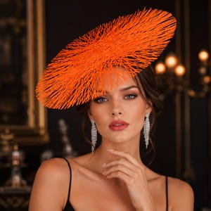Peut inclure: Un bibi orange vif présentant un large design circulaire en forme de disque composé de fines fibres de paille rayonnantes. La coiffe est associée à de longues boucles d'oreilles pendantes argentées composées de plusieurs rangées de cristaux.