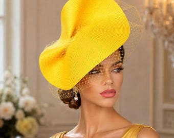 May include: A bright yellow fascinator hat featuring a wide, sculptural, curved brim made of woven straw material. The headpiece is adorned with a delicate, fine-mesh yellow netting veil that extends across the front, creating a textured overlay.