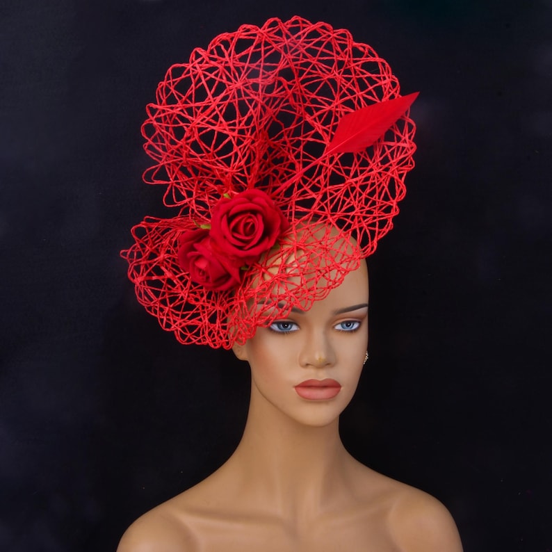 May include: A bright red fascinator hat featuring an intricate, web-like wire structure. The headpiece is adorned with two realistic red fabric roses and a single red feather accent, positioned on the side of the circular, open-work design.